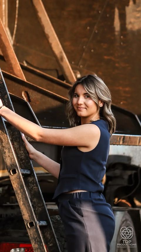 Close-up of a smiling woman in navy holding a truck ladder with a rusty tanker behind her