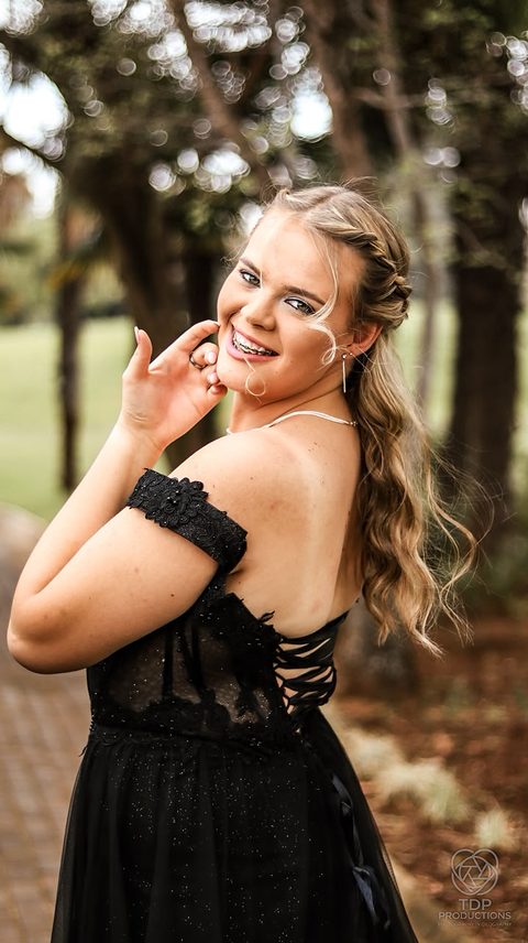 Young woman in a black off-the-shoulder gown smiling and touching her face among trees
