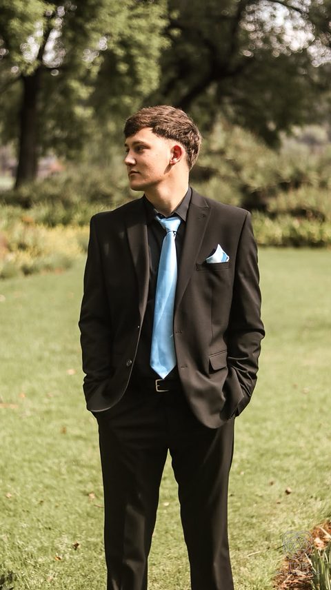 Young man in a dark suit with a light blue tie standing on a lawn with hands in pockets